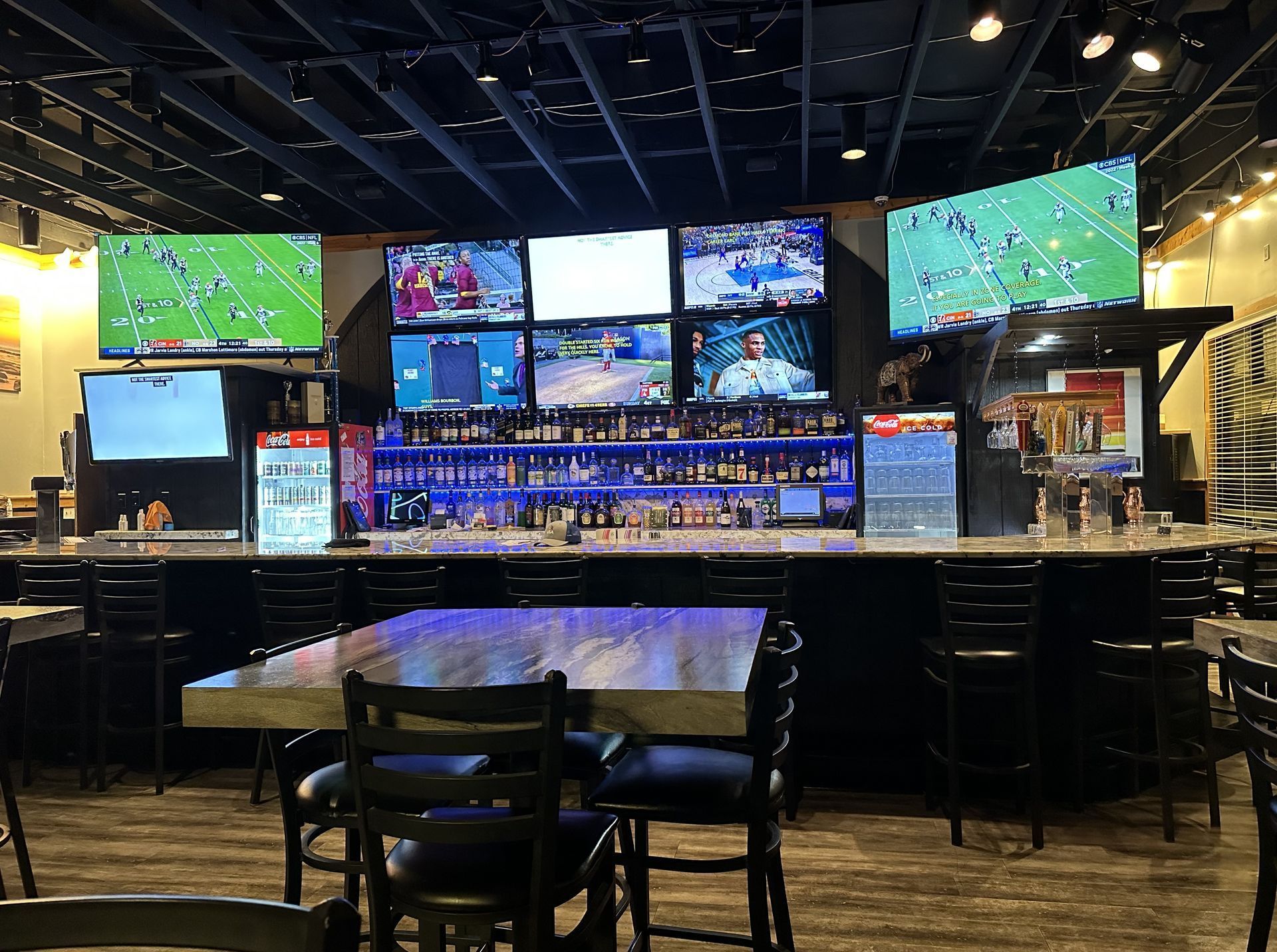 A bar with tables and chairs and a lot of televisions on the wall.