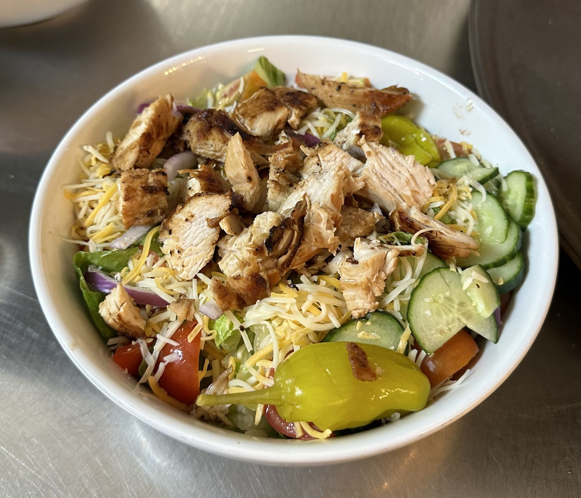 A bowl of salad with chicken , cheese , tomatoes , cucumbers and peppers on a table.
