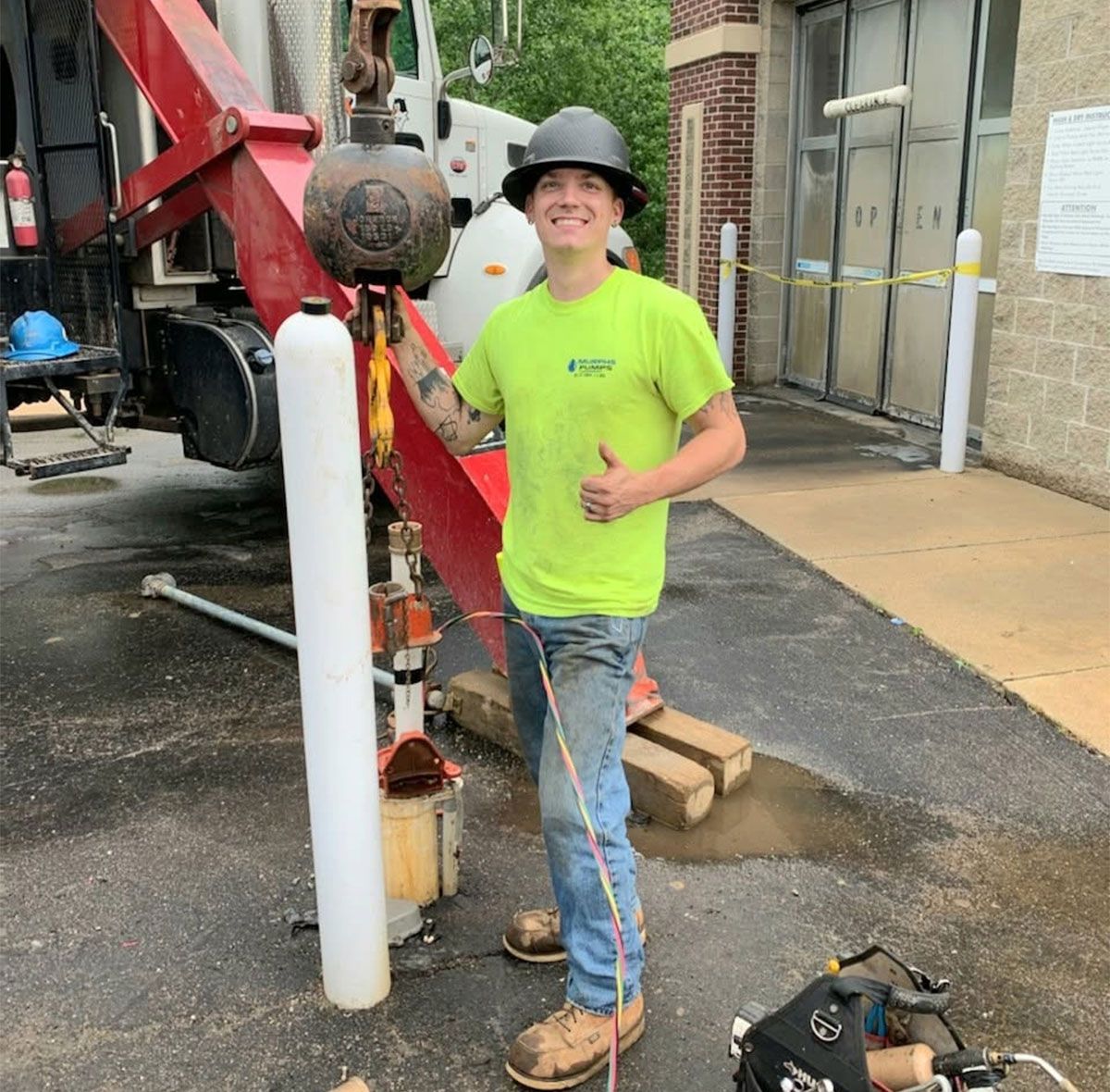 Worker in neon shirt and hard hat beside drilling equipment at a construction site, smiling and giving a thumbs-up
