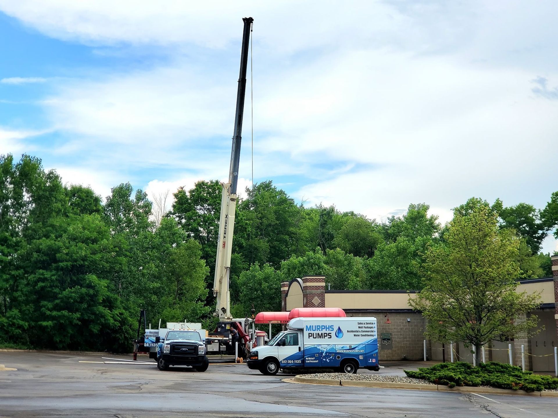 Crane lifting equipment beside a blue van and trucks in a parking lot near trees and a building