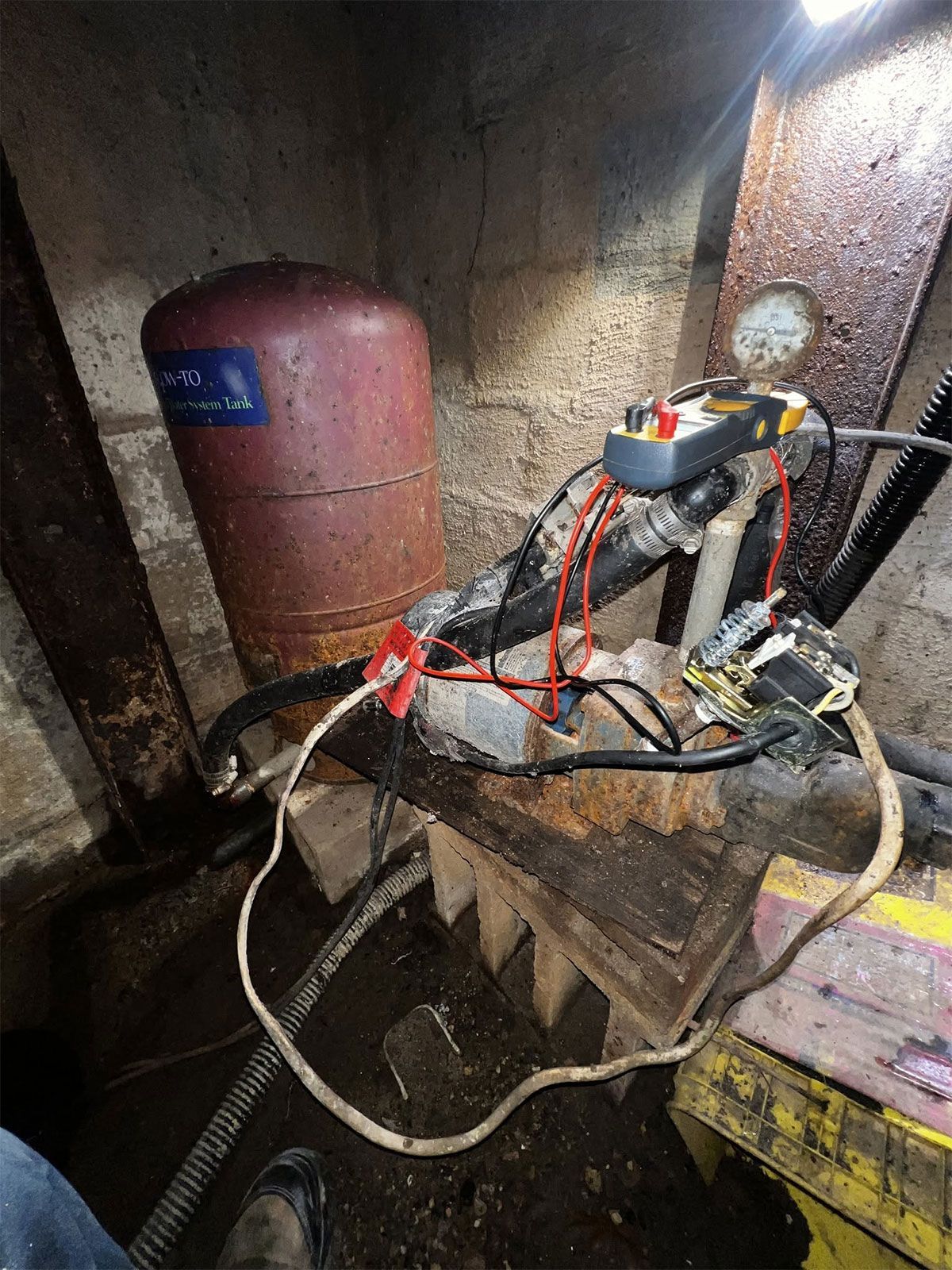 Dim utility space with red pressure tank, pipes, and tangled hoses on a rough wooden platform.