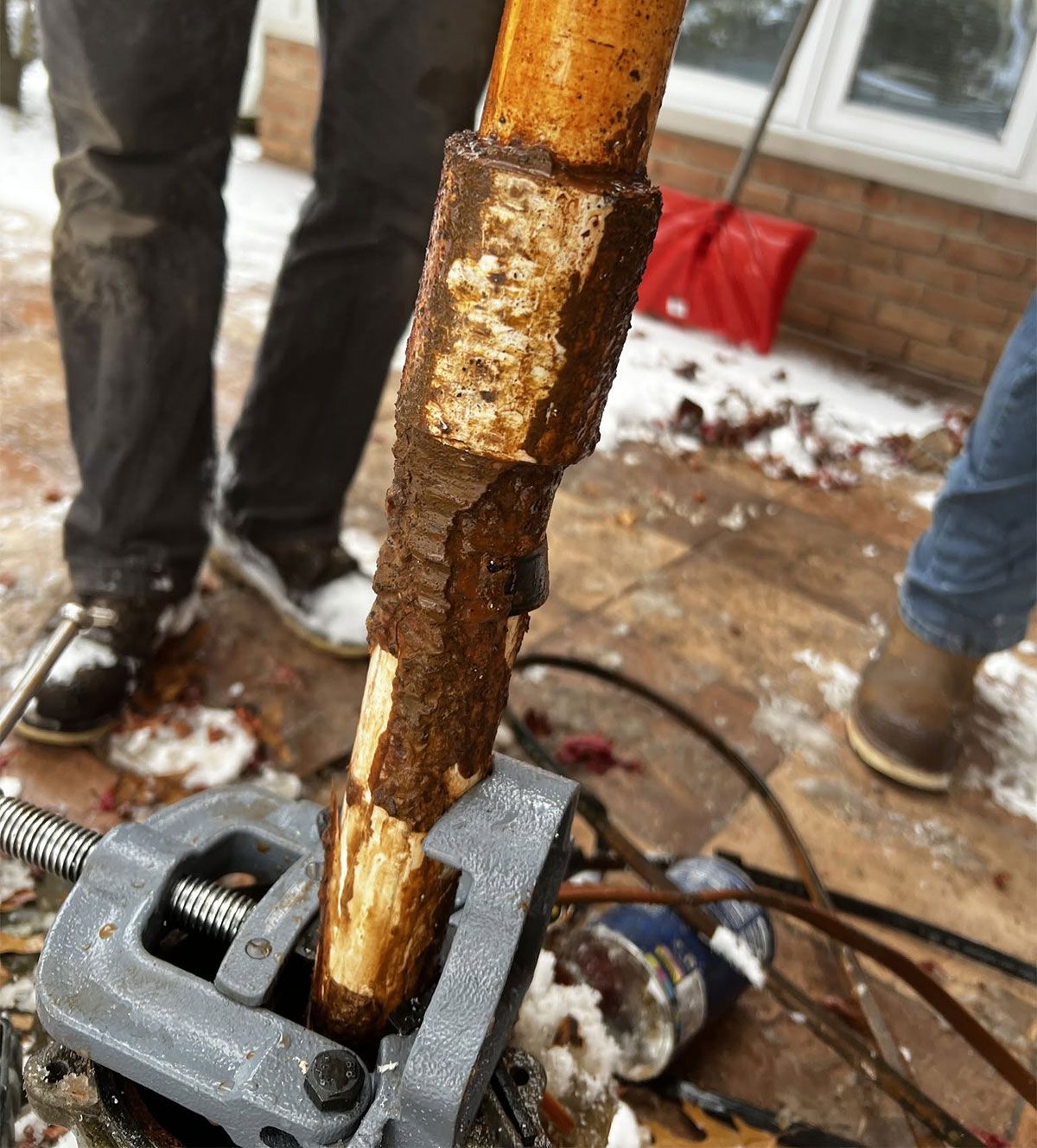 Rusty pipe being cut by a power tool outdoors in winter, with two people standing nearby.