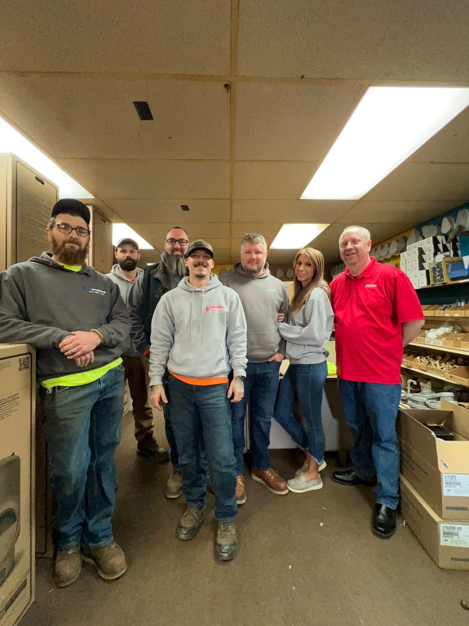 Group of six people standing in a store aisle, smiling beside stacked boxes and shelves.