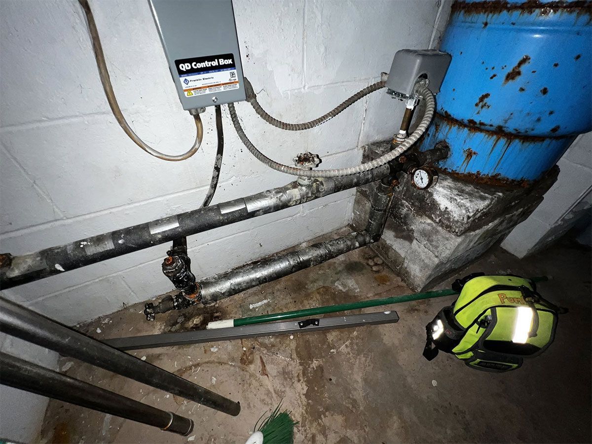 Basement utility area with blue tank, pipes, water on floor, and a flashlight near the wall.