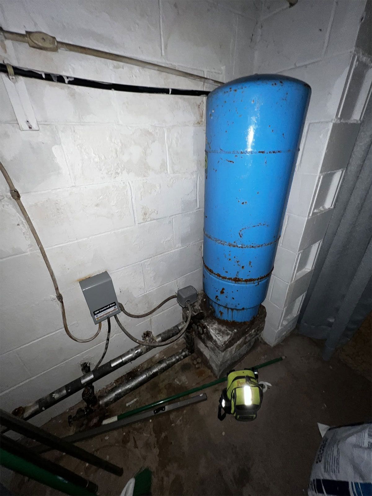 Basement utility area with blue tank, pipes, wall fixtures, and a small yellow tool on the floor