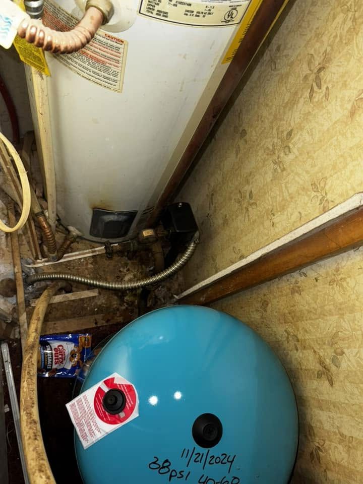 Large blue water heater in a cramped utility closet beside a white tank and exposed plumbing.