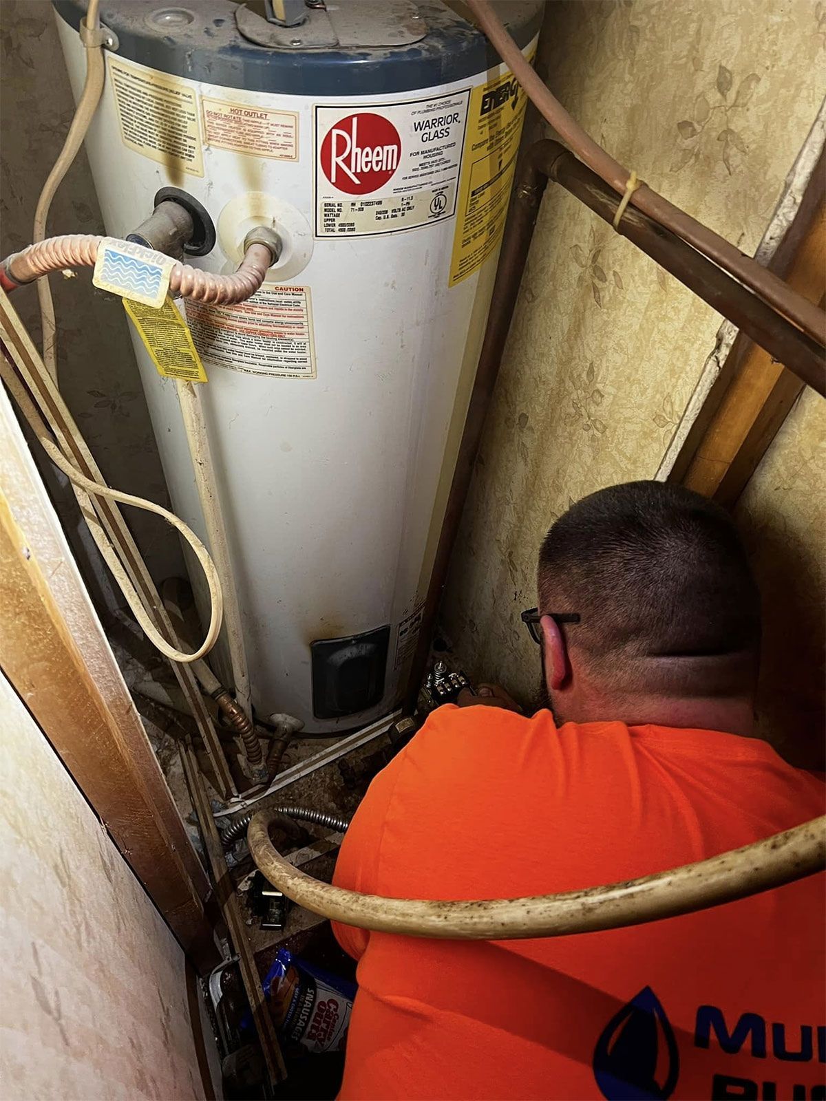 Worker crouches beside a water heater in a tight utility closet, inspecting pipes and fittings.