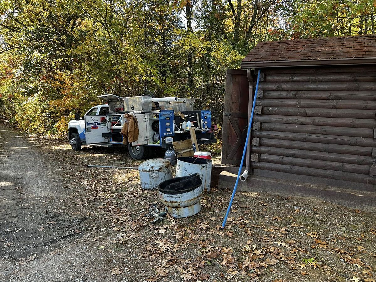 Rustic cabin beside a gravel yard with old vehicles, barrels, and autumn trees.