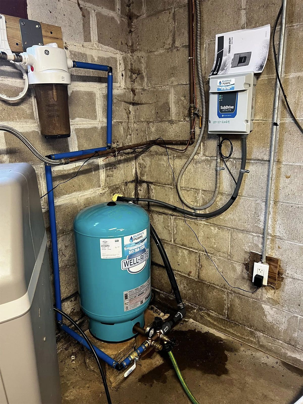 Basement utility corner with a blue pressure tank, pipes, and wall-mounted control box over a damp floor.