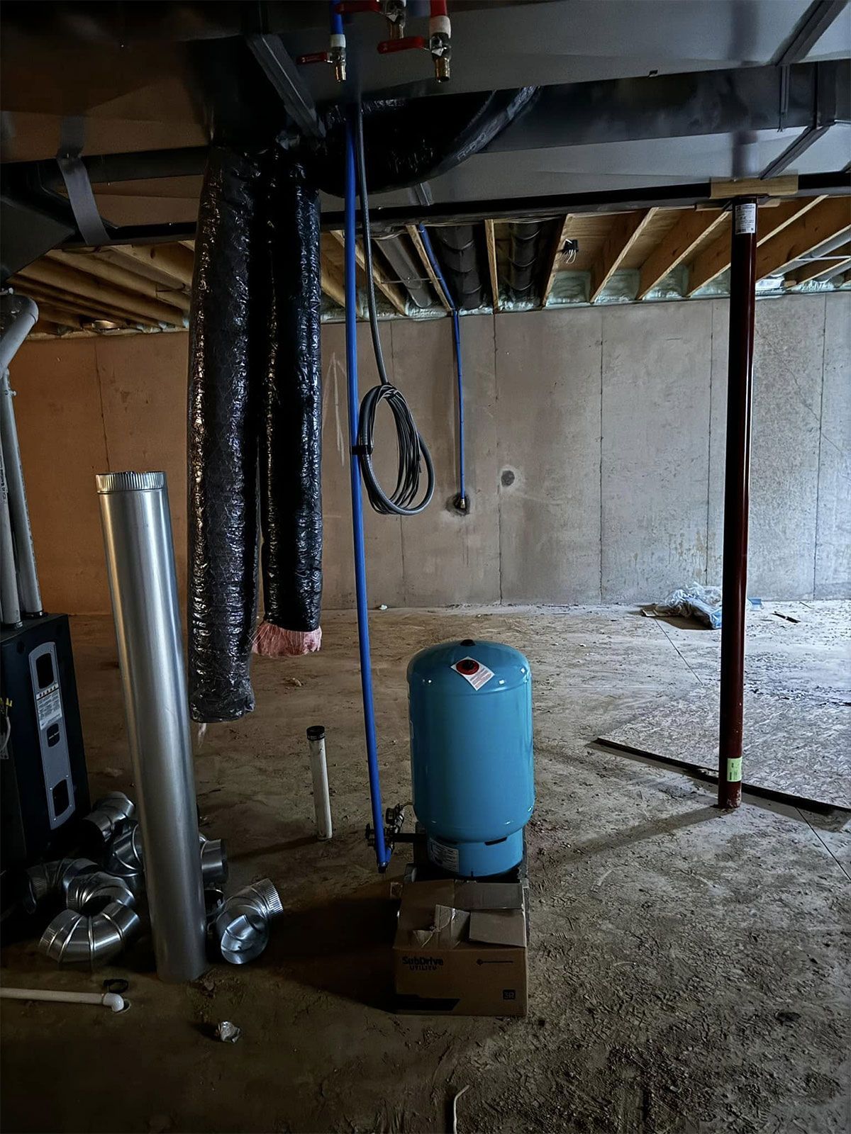 Unfinished basement with exposed joists, hanging cables, a blue tank, and construction materials on a dirt floor