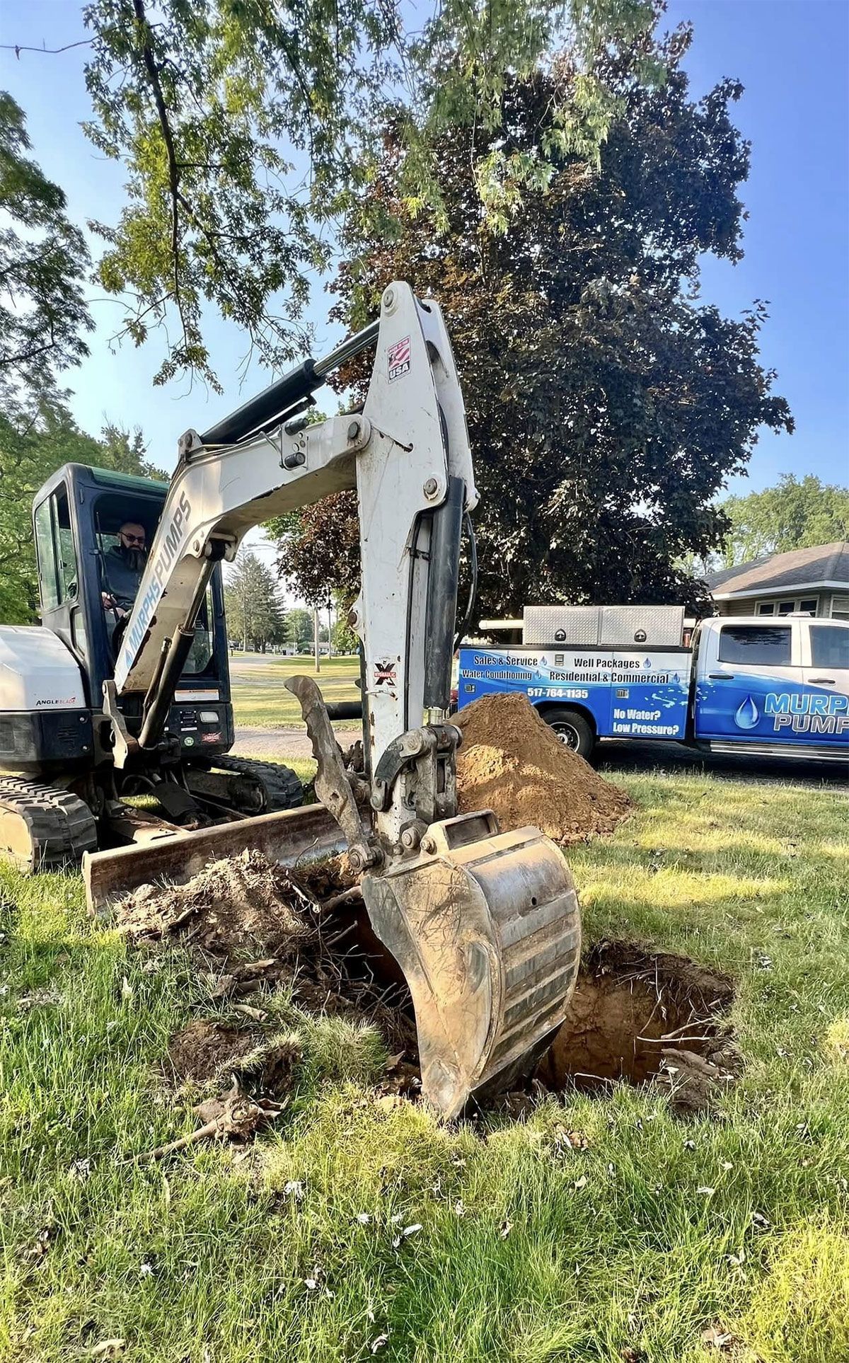 Excavator digging a hole in a grassy yard beside blue utility trucks and trees