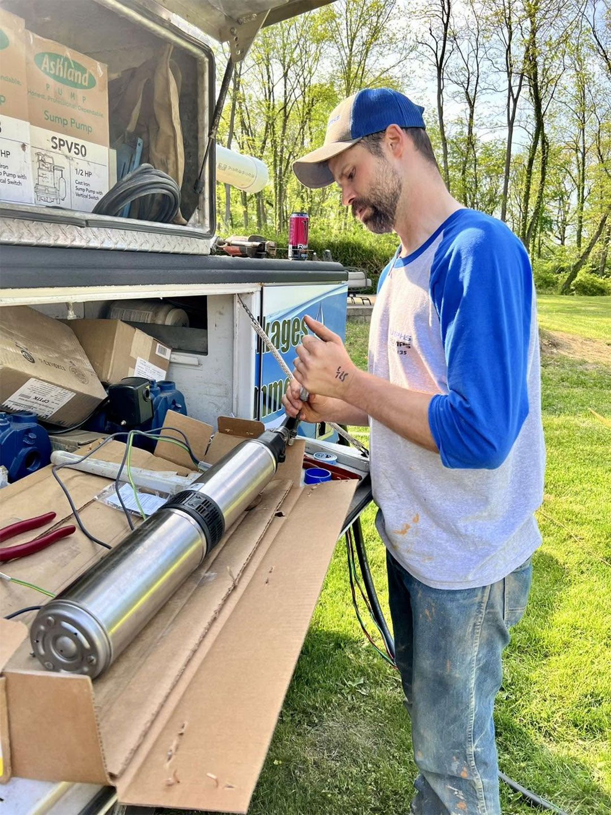 Man in blue cap and white shirt working on a metal cylinder at an outdoor workbench.