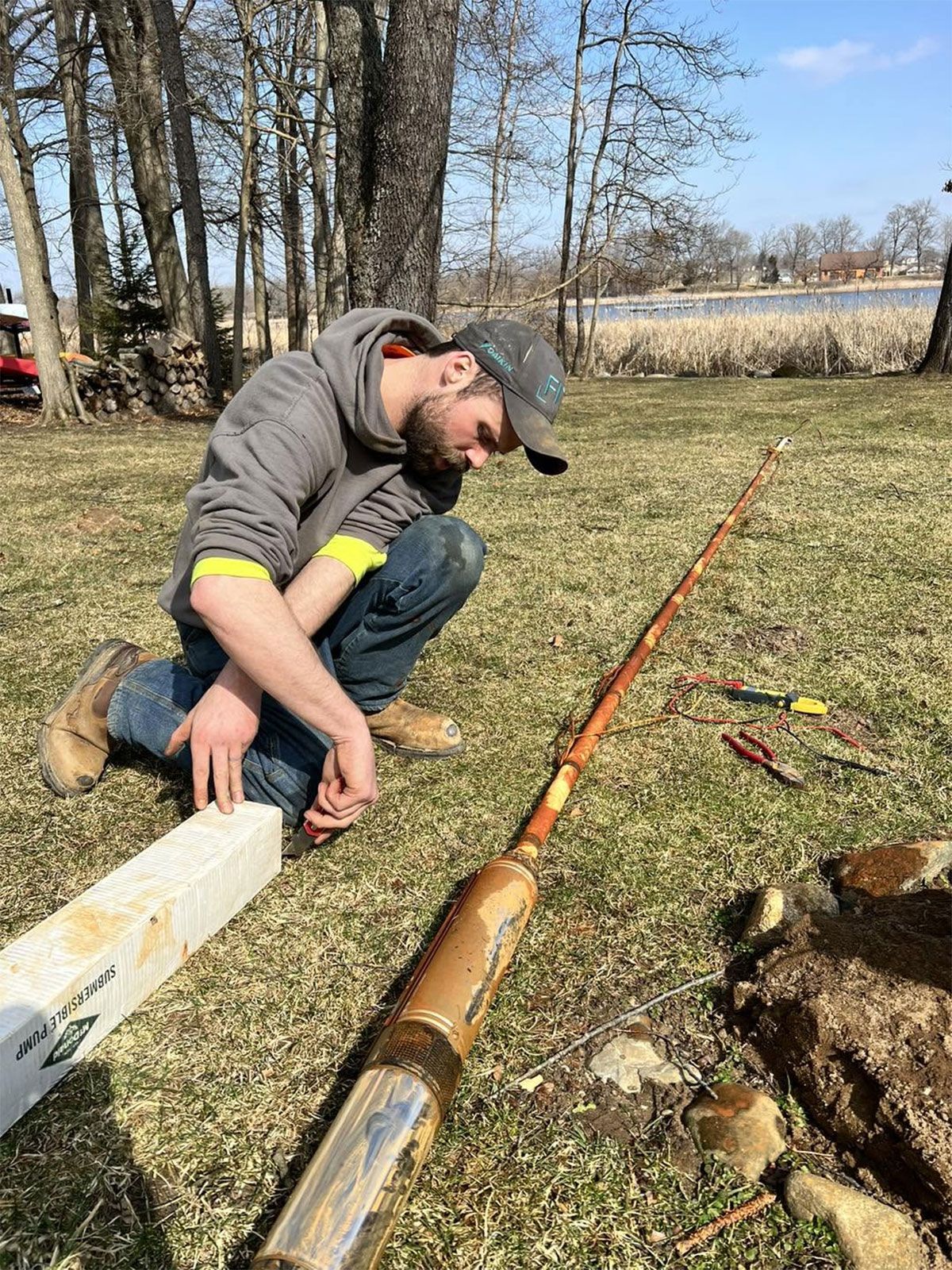 Man kneels outdoors beside a long pipe or log on a grassy field, inspecting it near trees.