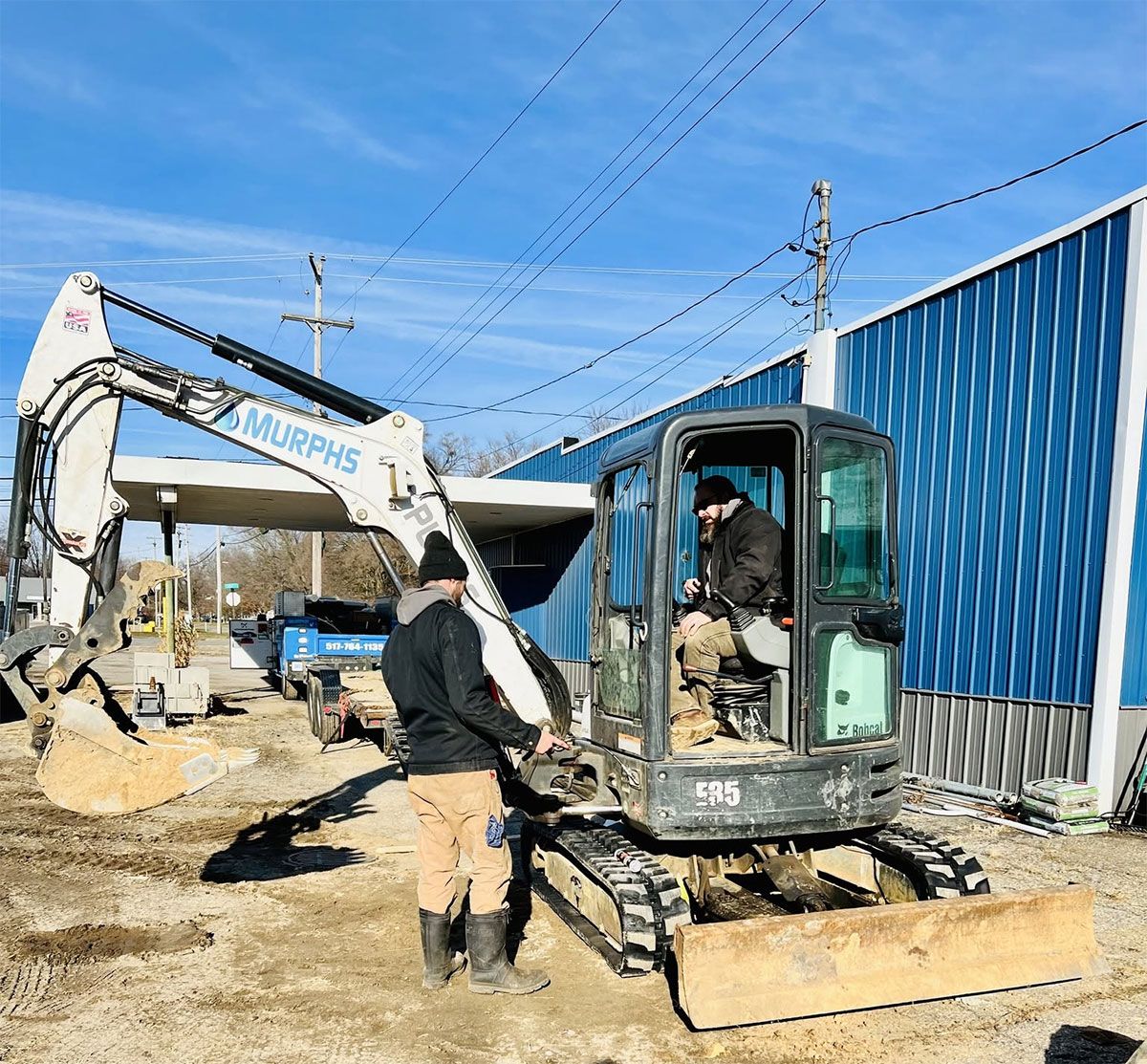 Two workers operate a compact excavator beside a blue metal building on a sunny construction site.