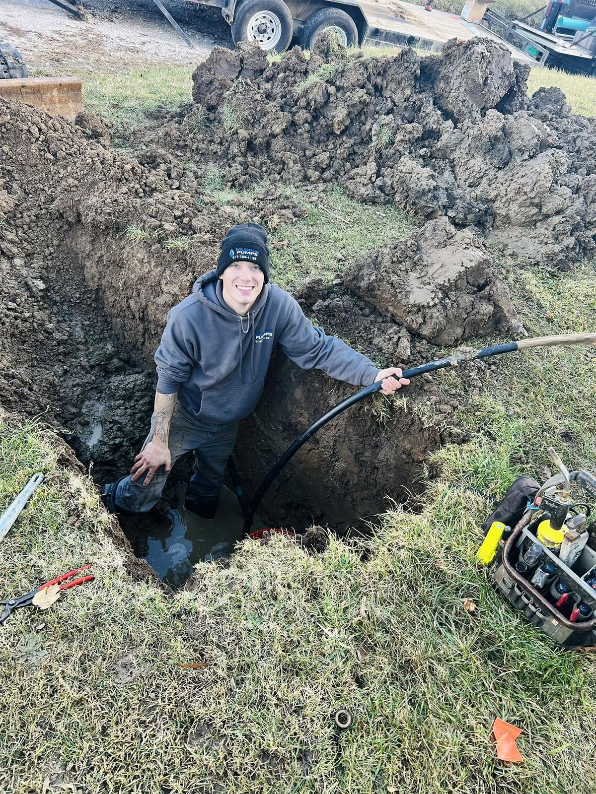 Worker standing in a deep dirt hole, holding a hose beside excavating equipment and soil piles outdoors