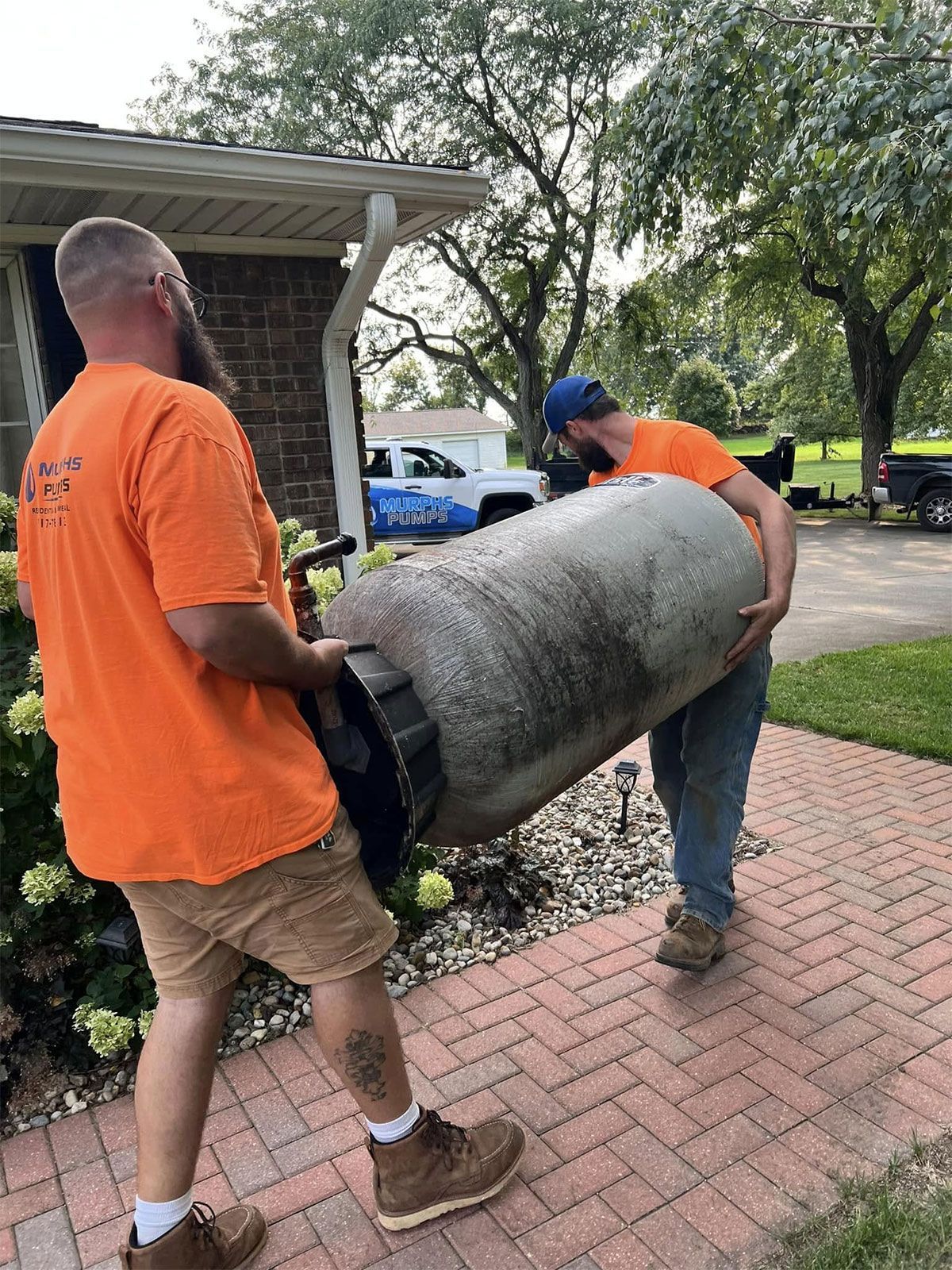 Two workers carrying a rolled gray carpet across a brick walkway near a house and parked truck.