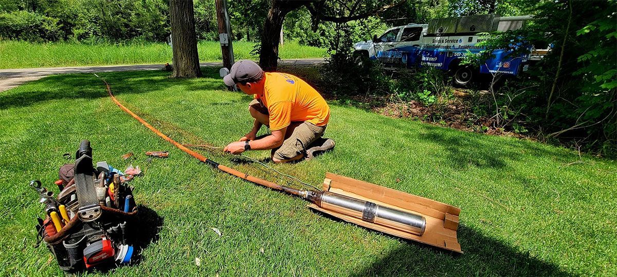 Worker in orange shirt kneeling on grass, using a tool beside a long wooden board and lawn equipment.