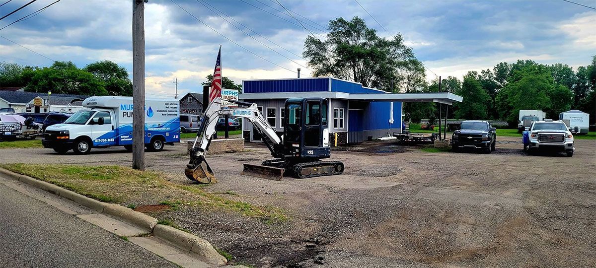 Roadside construction area with excavator, utility poles, and parked vehicles beside a blue building.