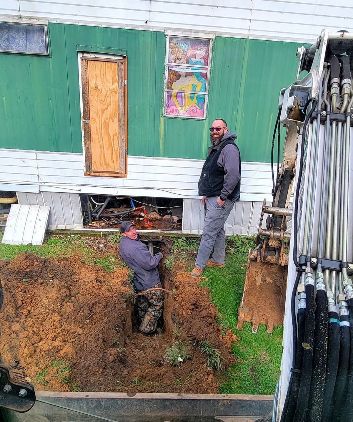 Two workers near a house foundation excavation beside green siding, one standing and one digging.