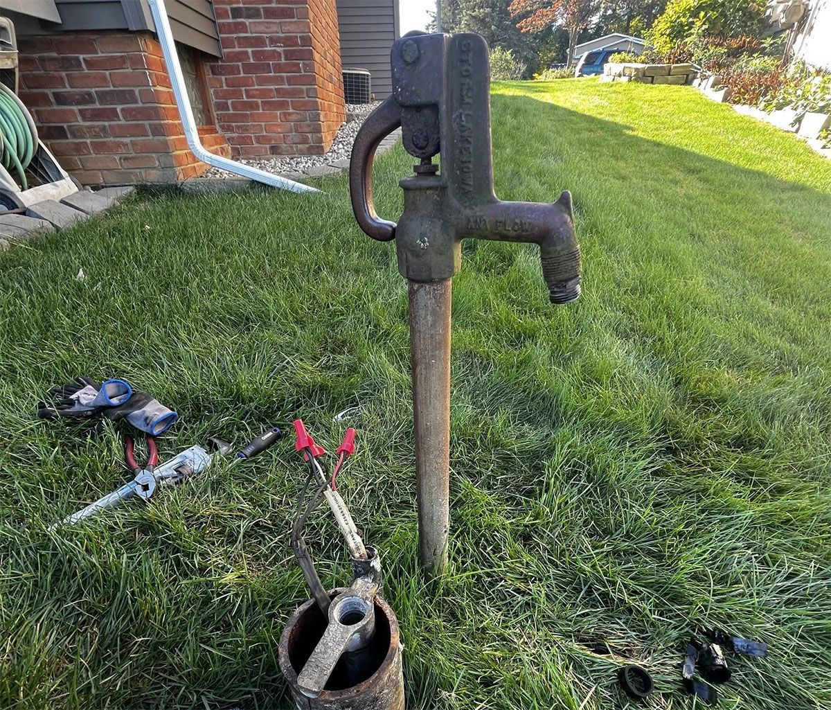 Old yard water pump with tools on grass beside a brick house