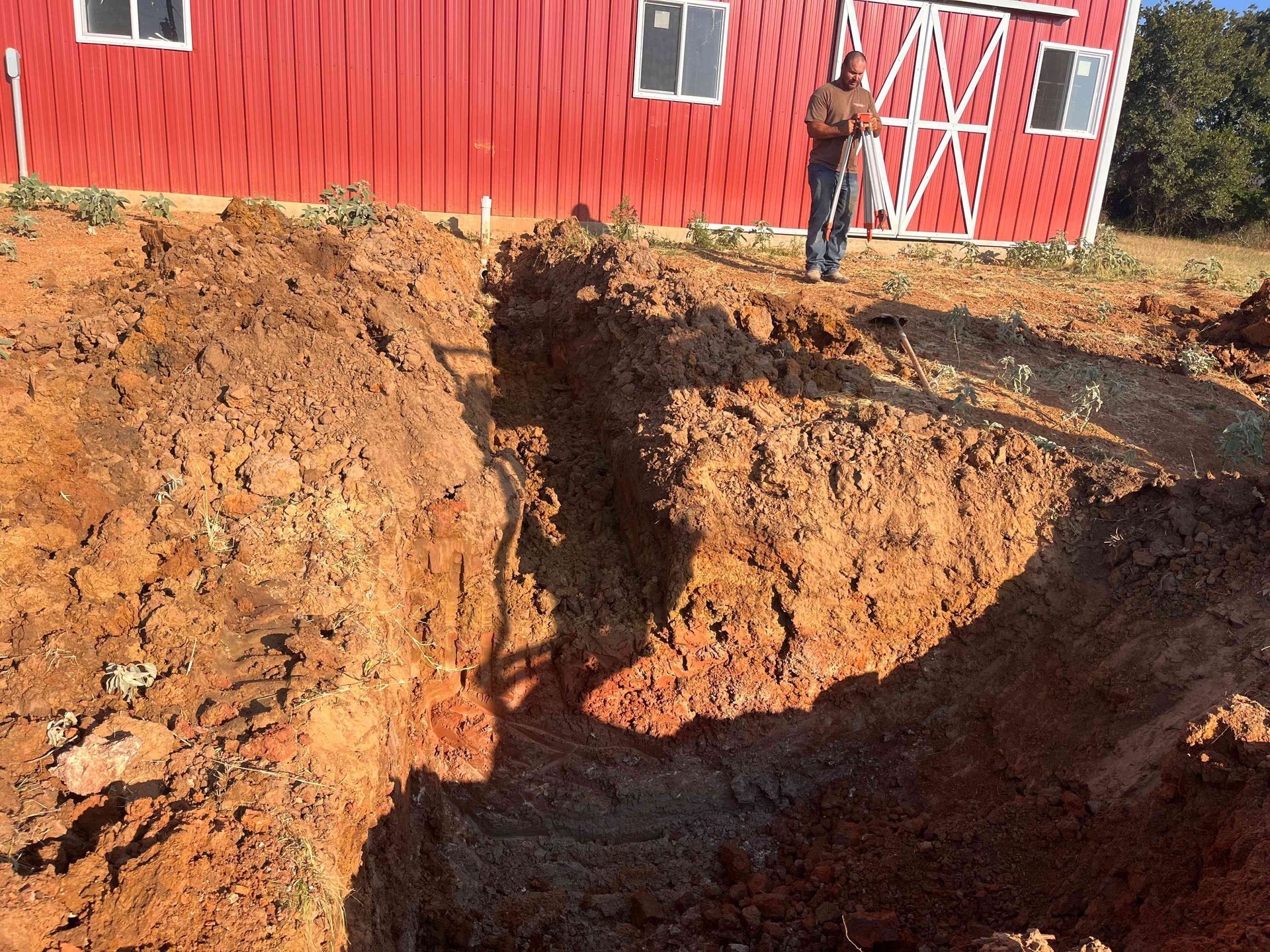 A person stands near a deep, narrow trench dug into reddish-brown soil in front of a bright red barn.