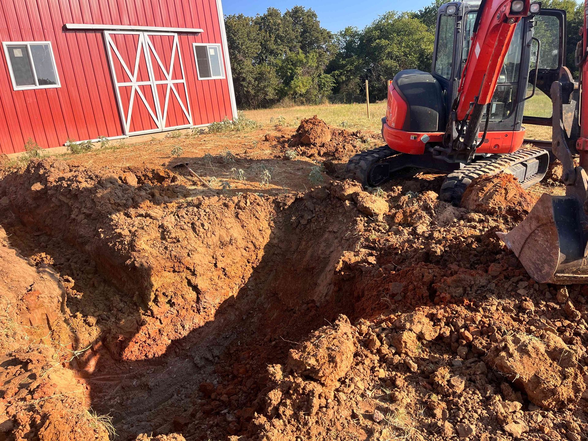 A red mini excavator sits next to a deep trench in the dirt in front of a red metal barn on a sunny day.