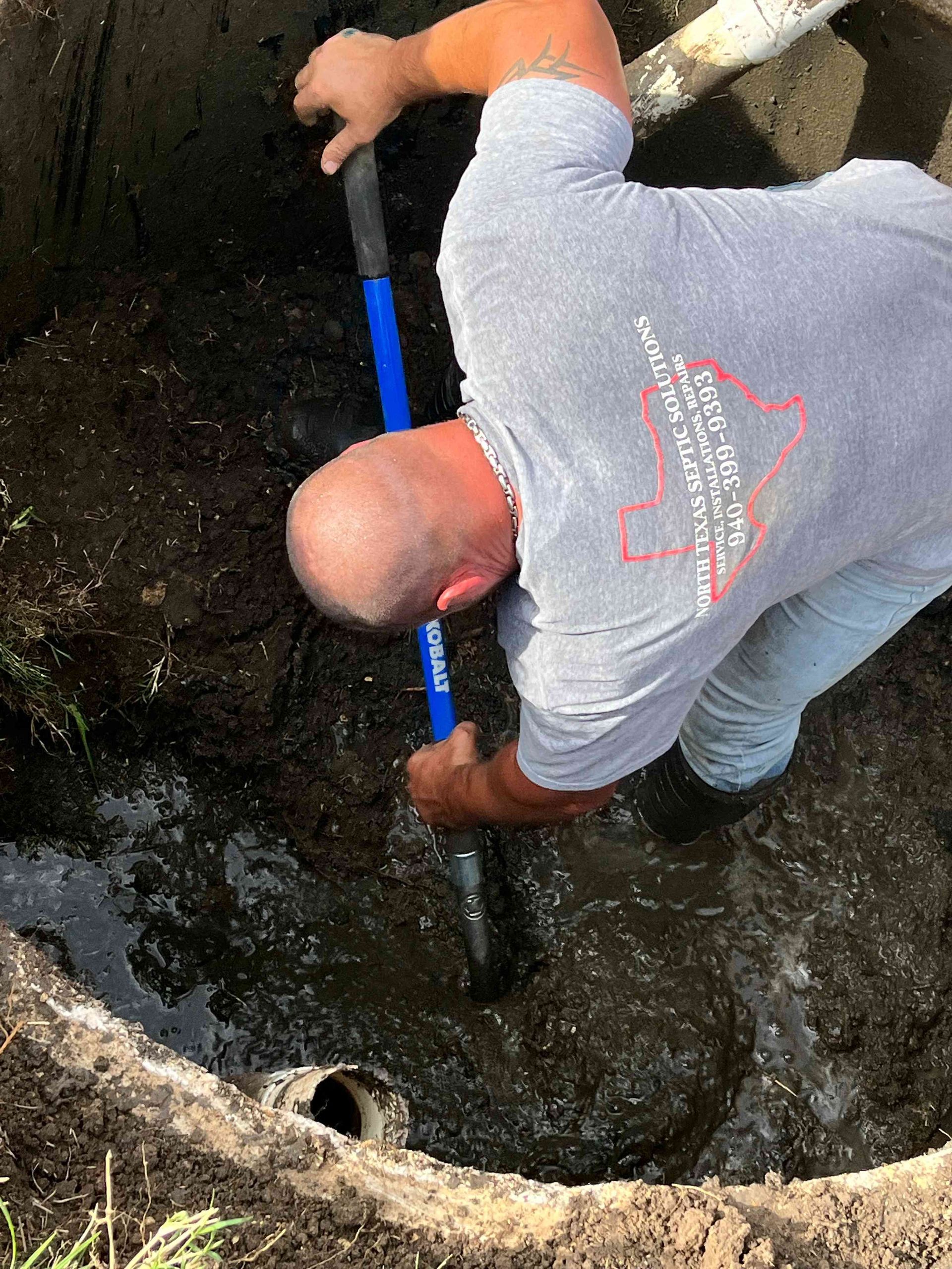 A worker wearing a gray t-shirt uses a blue shovel to dig in a muddy, circular hole in the ground.