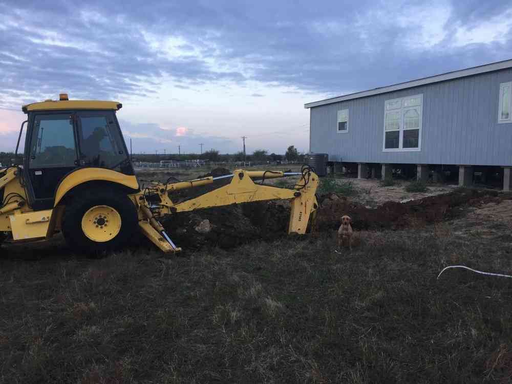 A yellow backhoe digs a trench next to a raised gray house at dusk, with a small dog standing nearby.