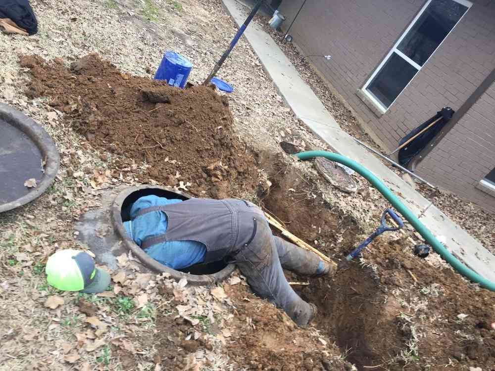 A person wearing work clothes leans into an open, in-ground septic tank in a yard, with soil piled nearby.