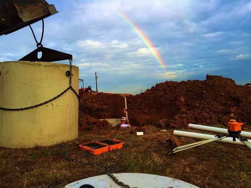 A construction site with a large concrete pipe section, mounds of dirt, equipment, and a rainbow in the cloudy sky.