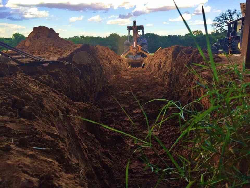 A freshly dug trench in a dirt field with a construction vehicle working in the distance under a cloudy sky.
