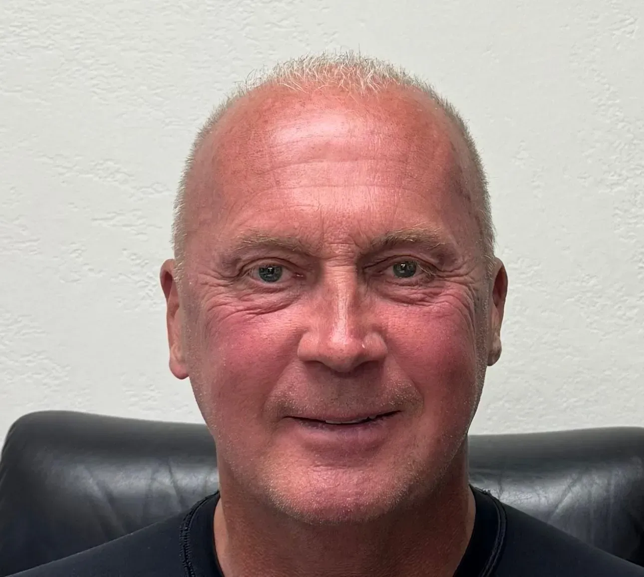 Smiling person seated against a white wall, wearing a black shirt.
