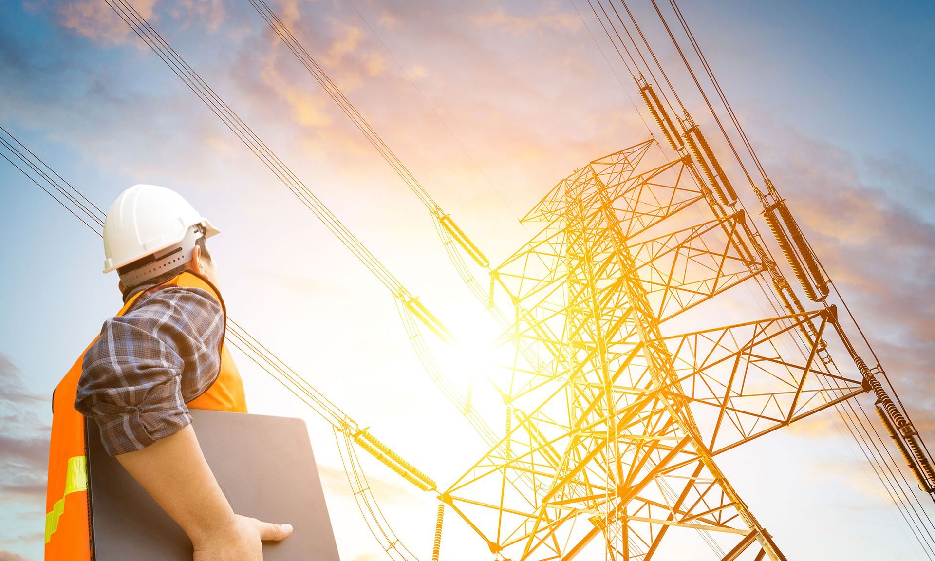 Worker in hard hat stands by power lines and transmission tower at sunrise.