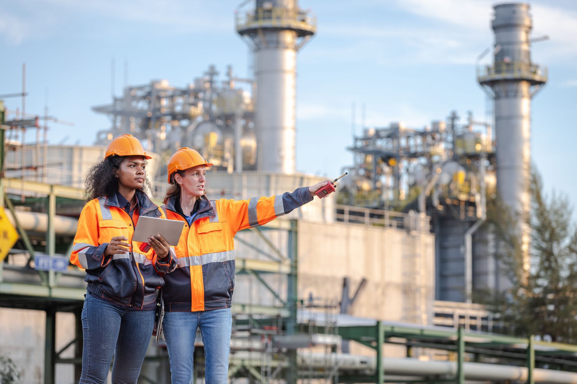 Two industrial workers in orange safety gear pointing at equipment at a refinery