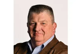 Portrait of a man in a brown jacket and blue shirt, smiling against a plain white background