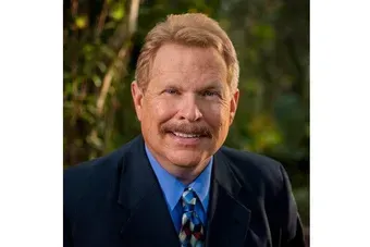 Portrait of a smiling man in a dark suit and blue shirt against a leafy outdoor background