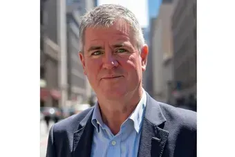 Man in a blue blazer stands on a city street with blurred buildings behind him