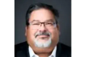 Smiling man with glasses and a white goatee in a dark blazer, against a plain background