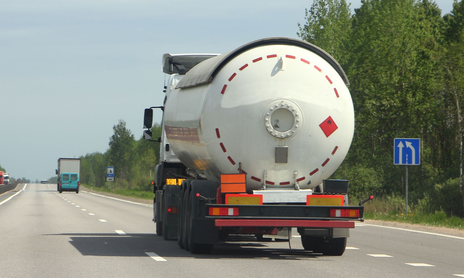 White tanker truck driving on a highway beside trees, viewed from behind.