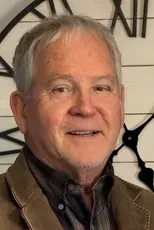 Smiling man in a brown jacket standing indoors against a decorative wall clock backdrop