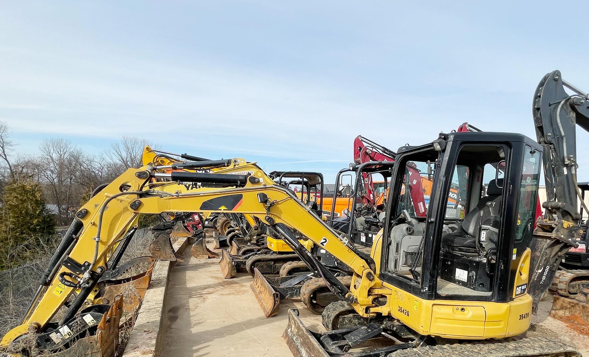 A row of yellow excavators are parked on a dirt road.
