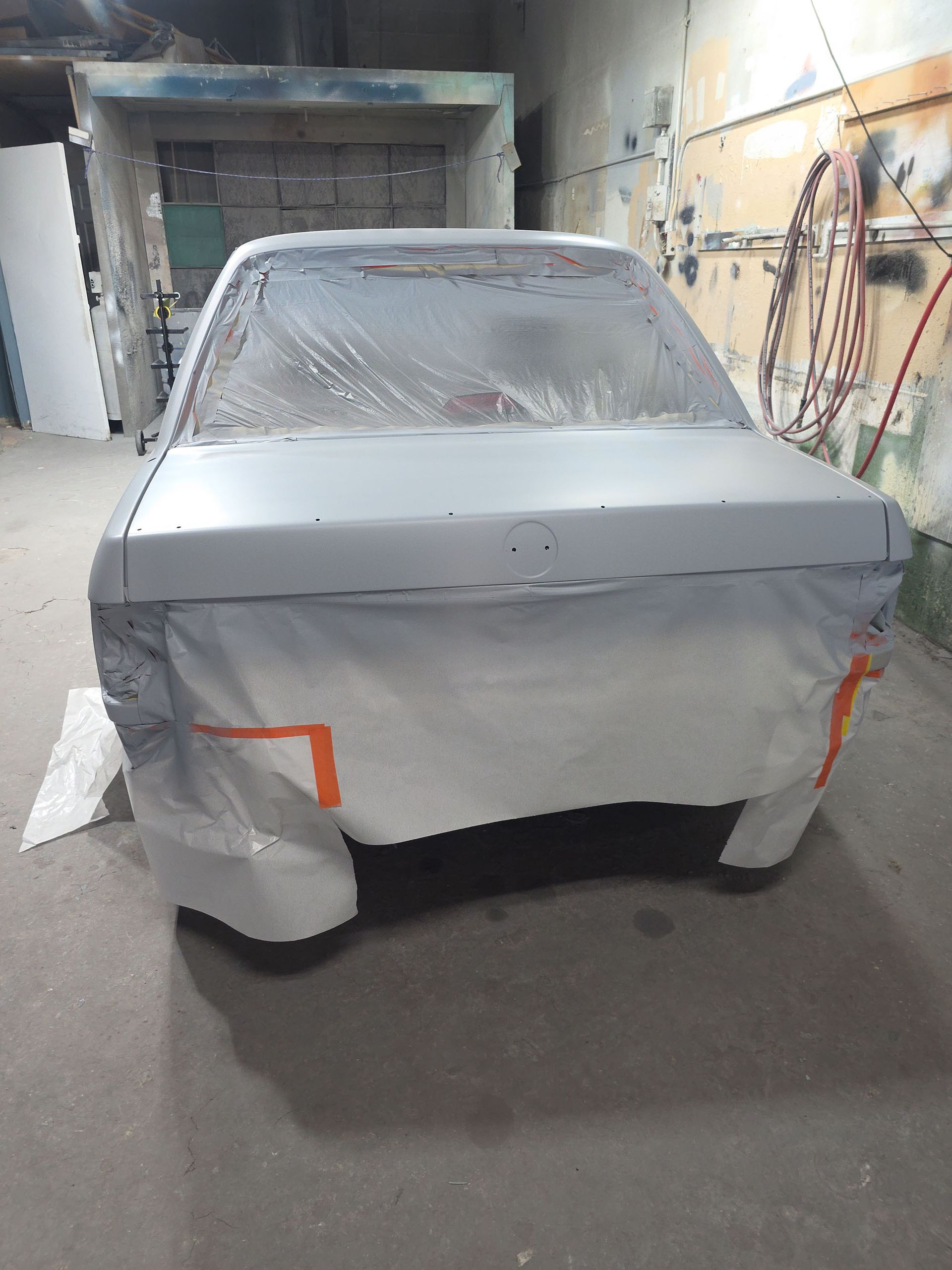 Car masked with gray plastic in a paint booth, front view, partially sanded and taped off.