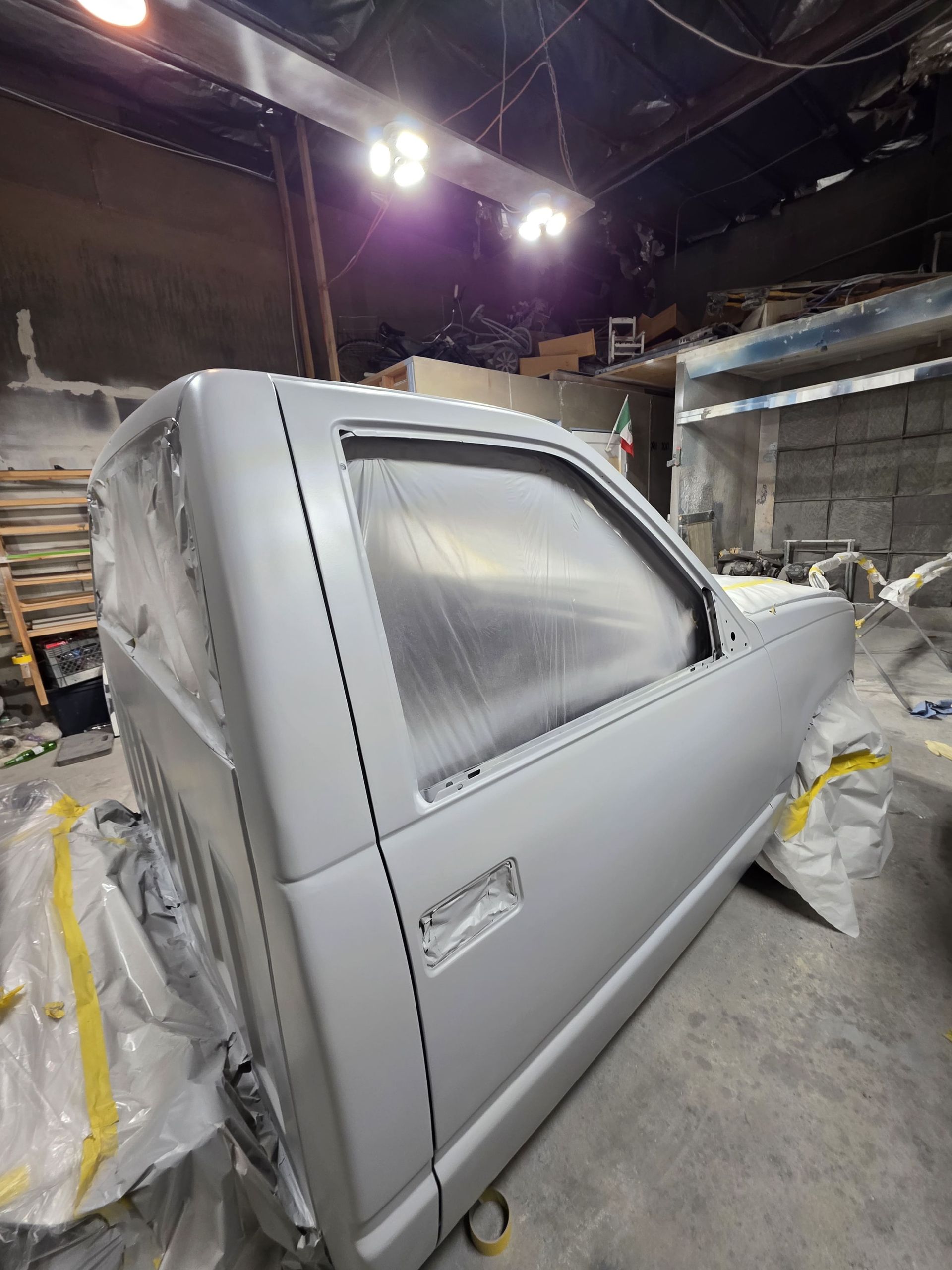 Gray pickup truck body in a workshop, covered in plastic with bright overhead lights.