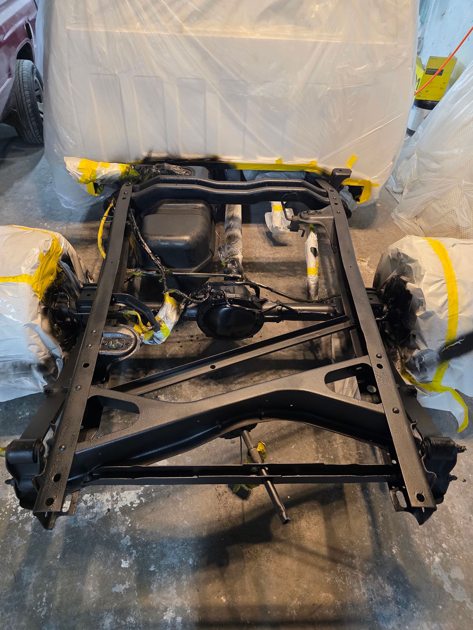 Bare car chassis frame with exposed engine, wrapped in clear plastic and yellow tape in a workshop.
