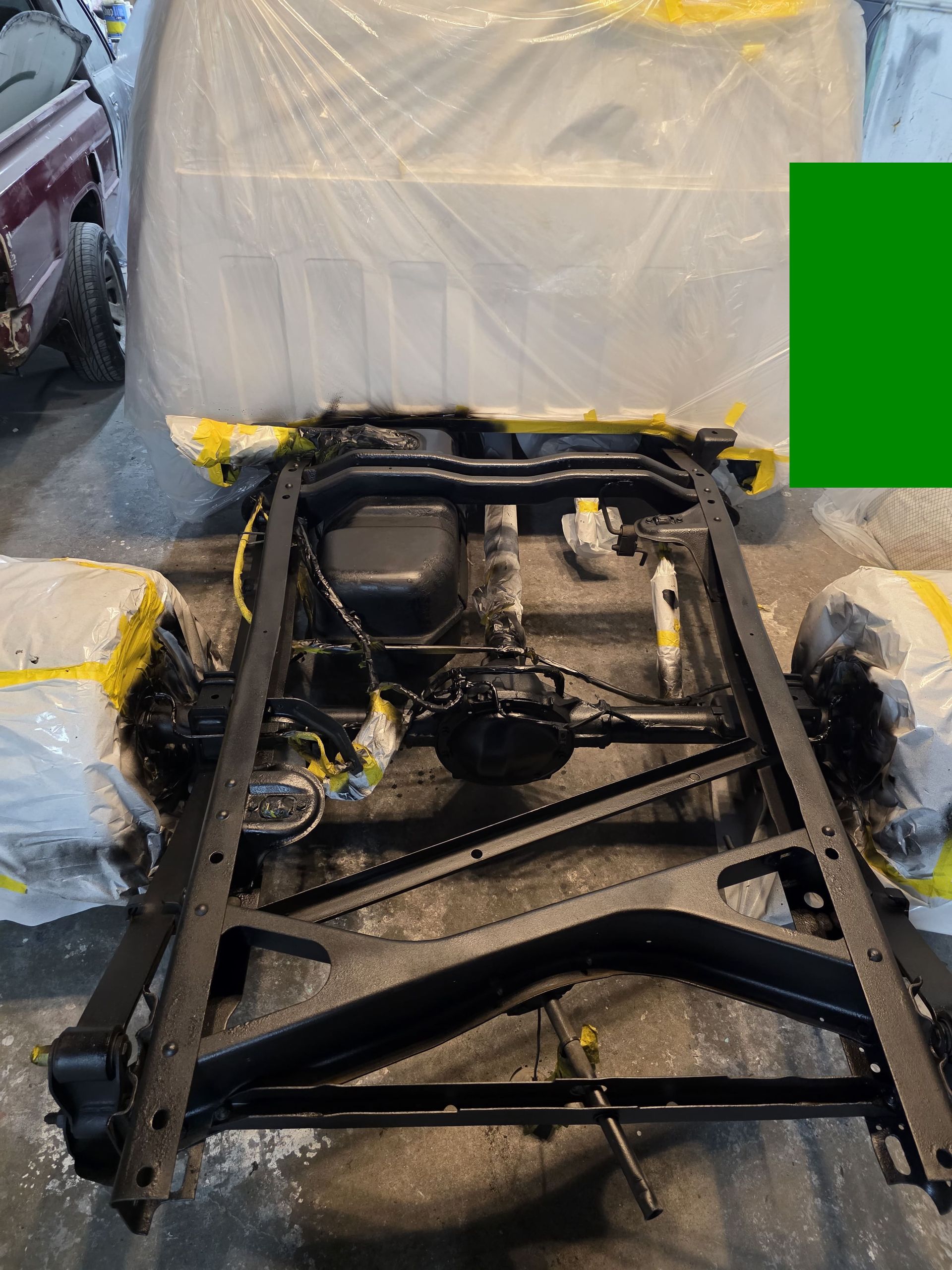 Black metal car chassis frame in a workshop, with a covered seat and parts wrapped in yellow tape.