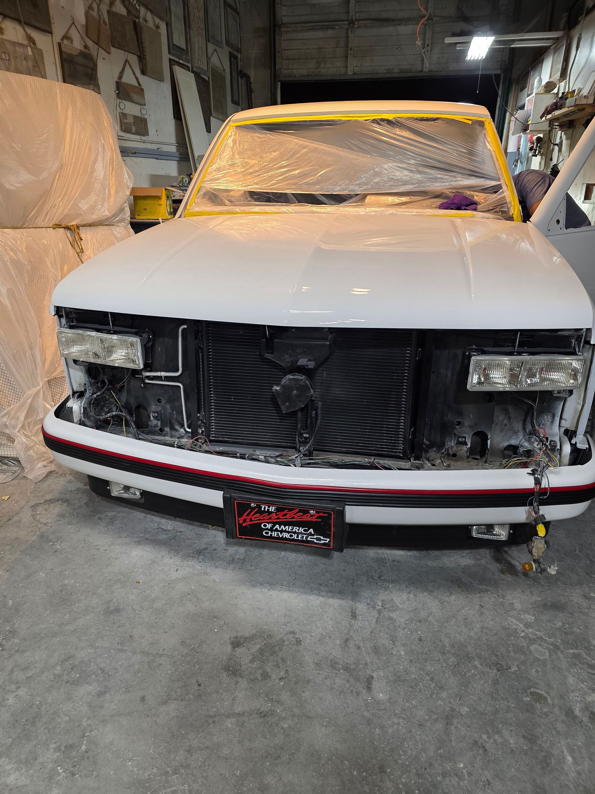 Damaged white car in a garage, front grille removed and hood dust-covered, with a red bumper stripe.