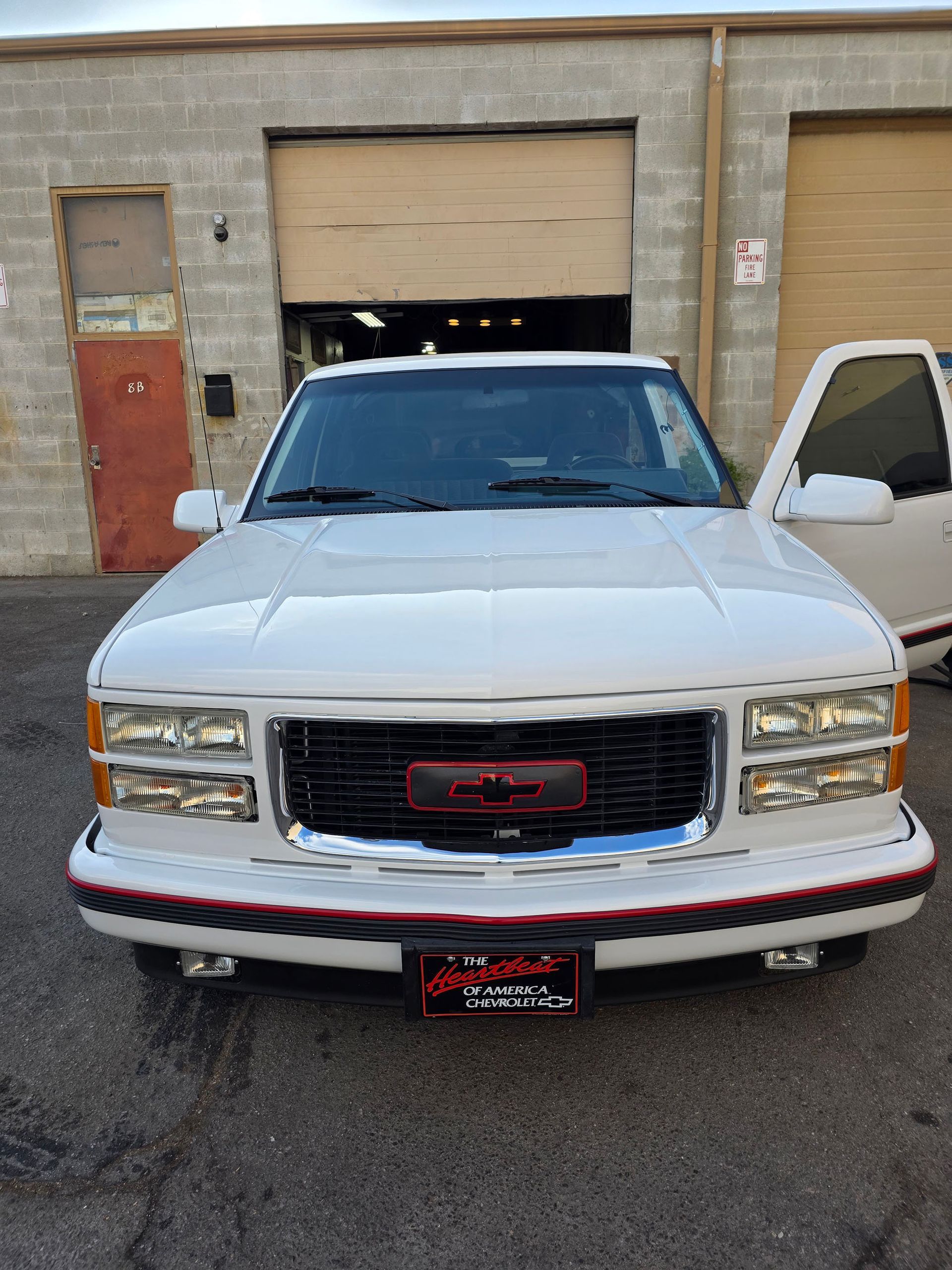 White GMC pickup truck parked in front of a warehouse, with the driver door open.