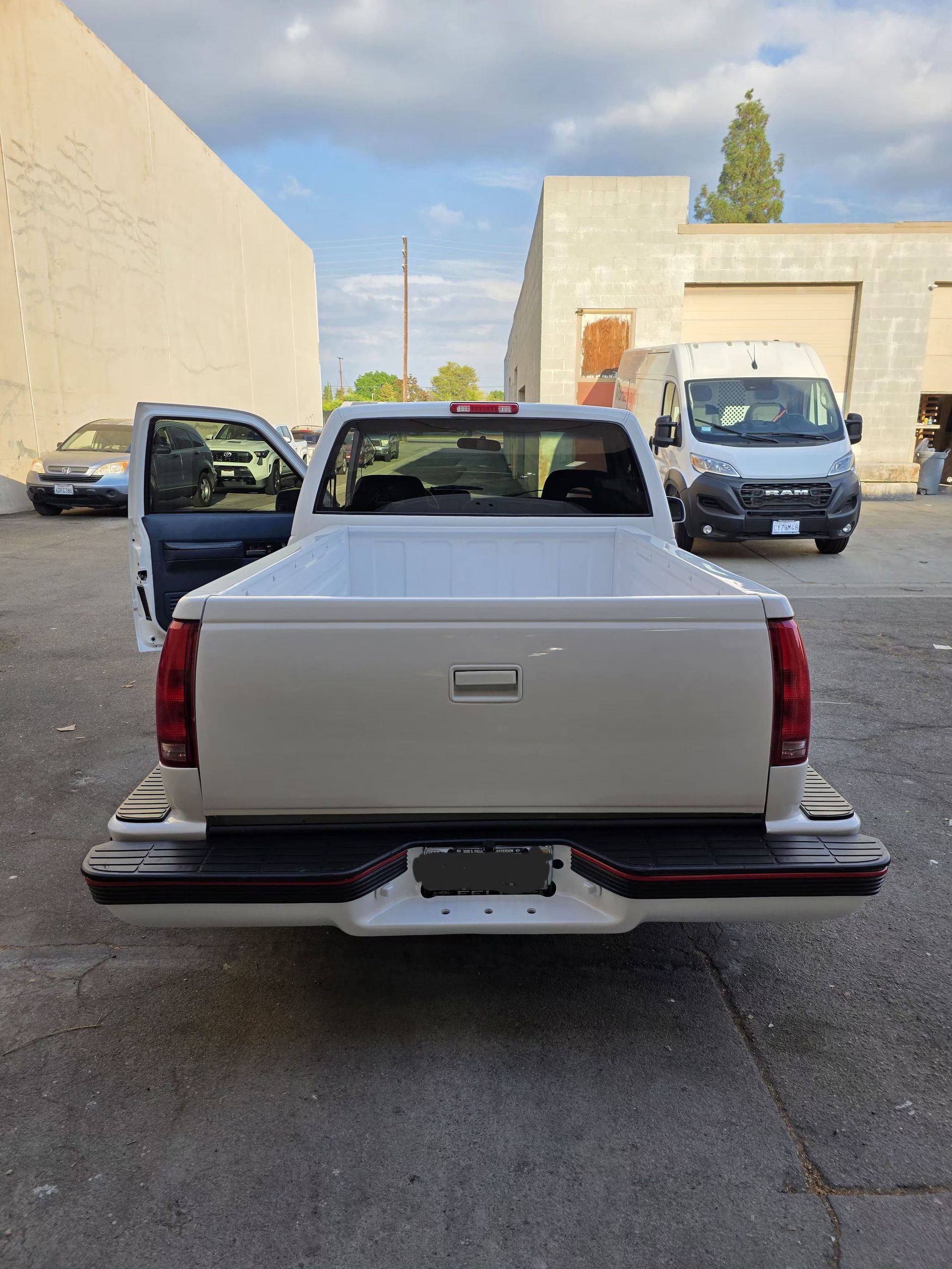 White pickup truck viewed from behind in an alley, with the driver door open and a van parked ahead.