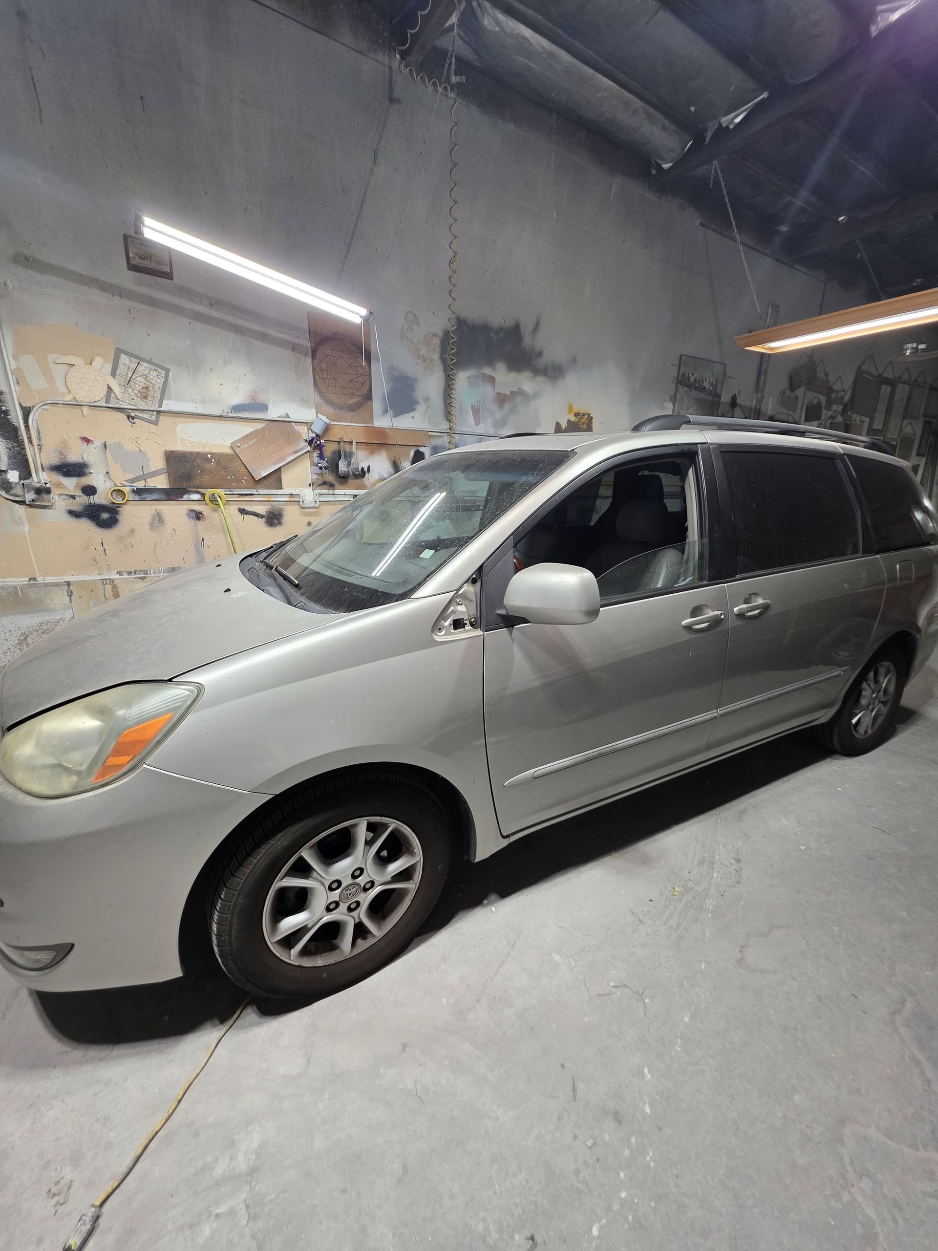 Silver minivan parked inside a garage or workshop with fluorescent lights overhead