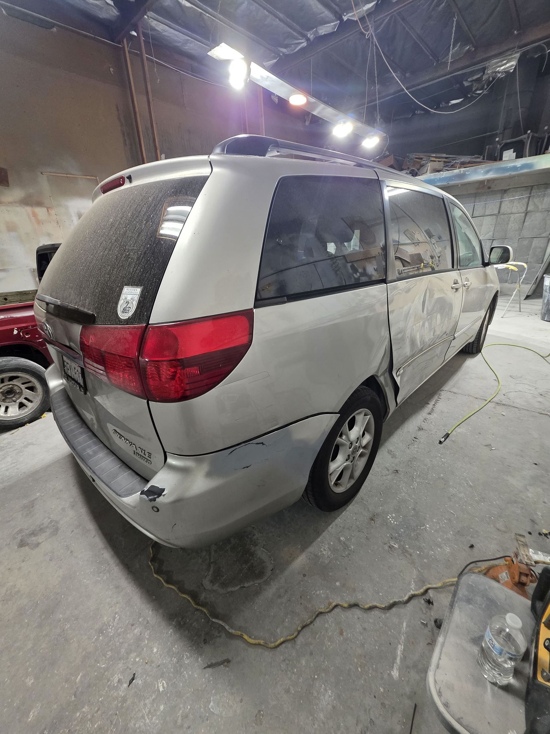 Silver minivan parked indoors in a garage, rear-left view, with visible body damage and dust.